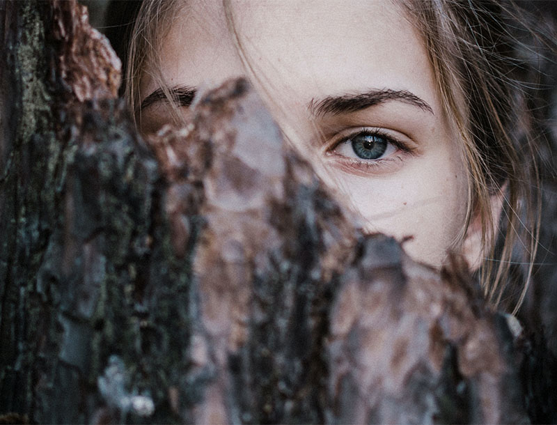Nahaufnahme eines blauen Auges einer Frau, das hinter einem Baumstamm hervorschaut.